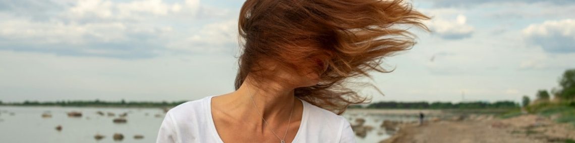 "A young woman with her hair blowing in the wind stands on the rocky shore of Kopli, Tallinn, looking out at the Baltic Sea under a dramatic cloudy sky."